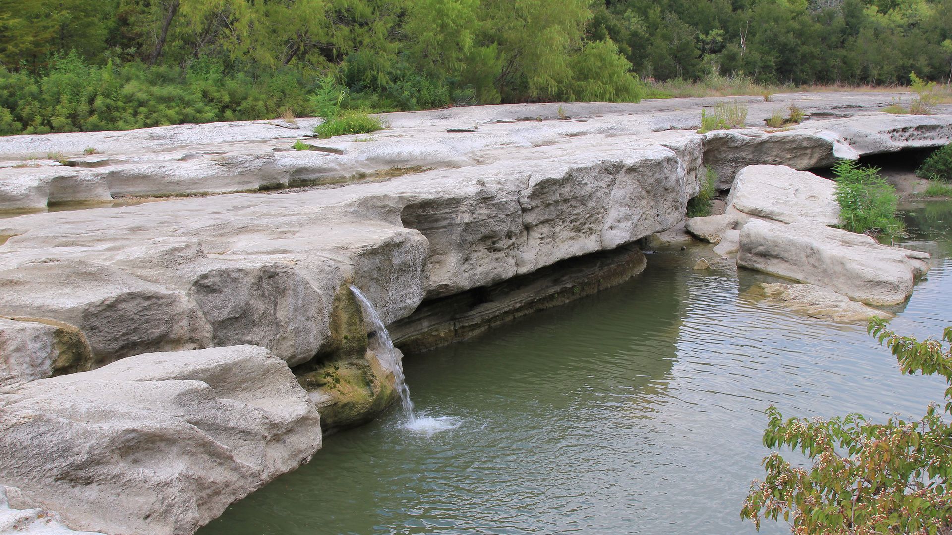 Water flows over a rock formation into a body of water, with green foliage in the background.