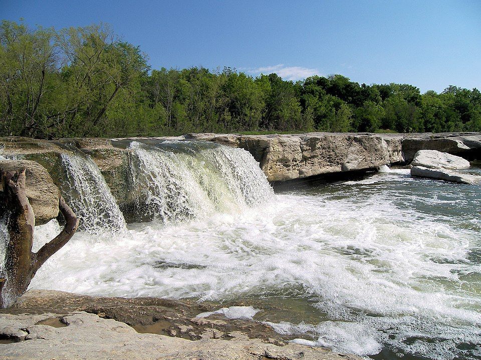 Waterfall cascading over layered stone, with foamy water, and trees in the background on a sunny day.
