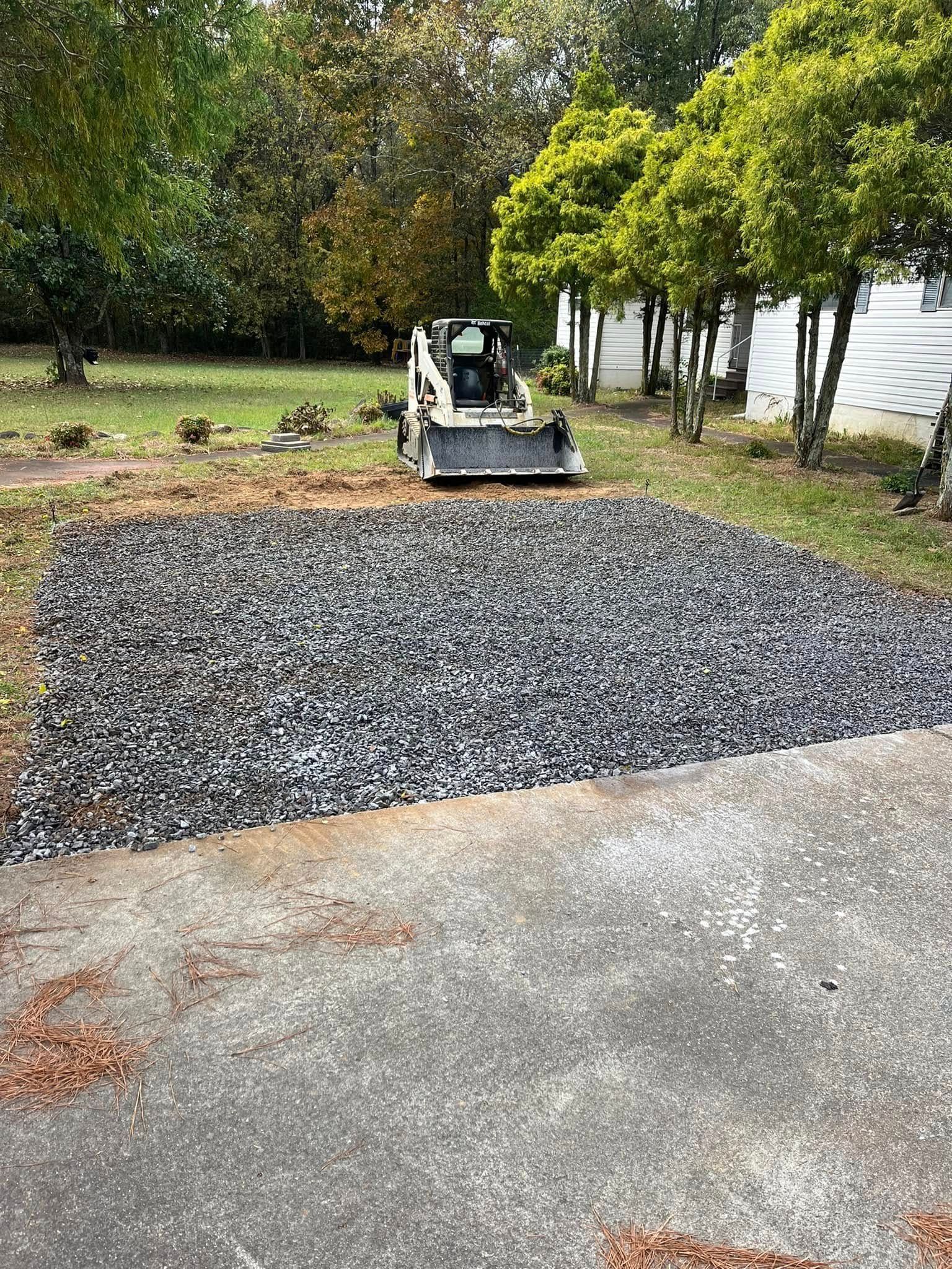 Gray gravel pile on a driveway beside a small excavator, with trees and a white building in the background