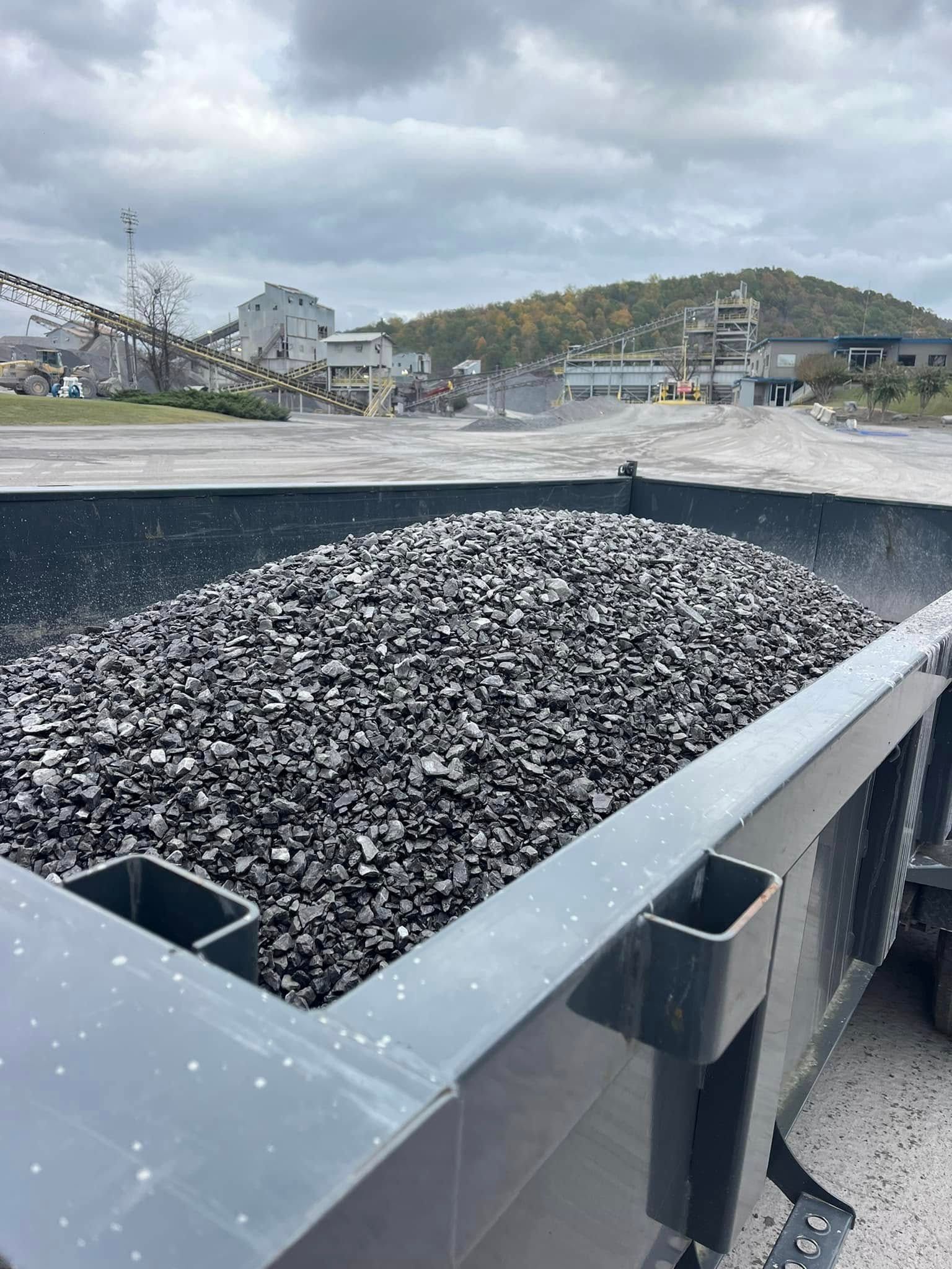 Truck bed filled with crushed black rock at a quarry, with cloudy sky and industrial equipment in the background