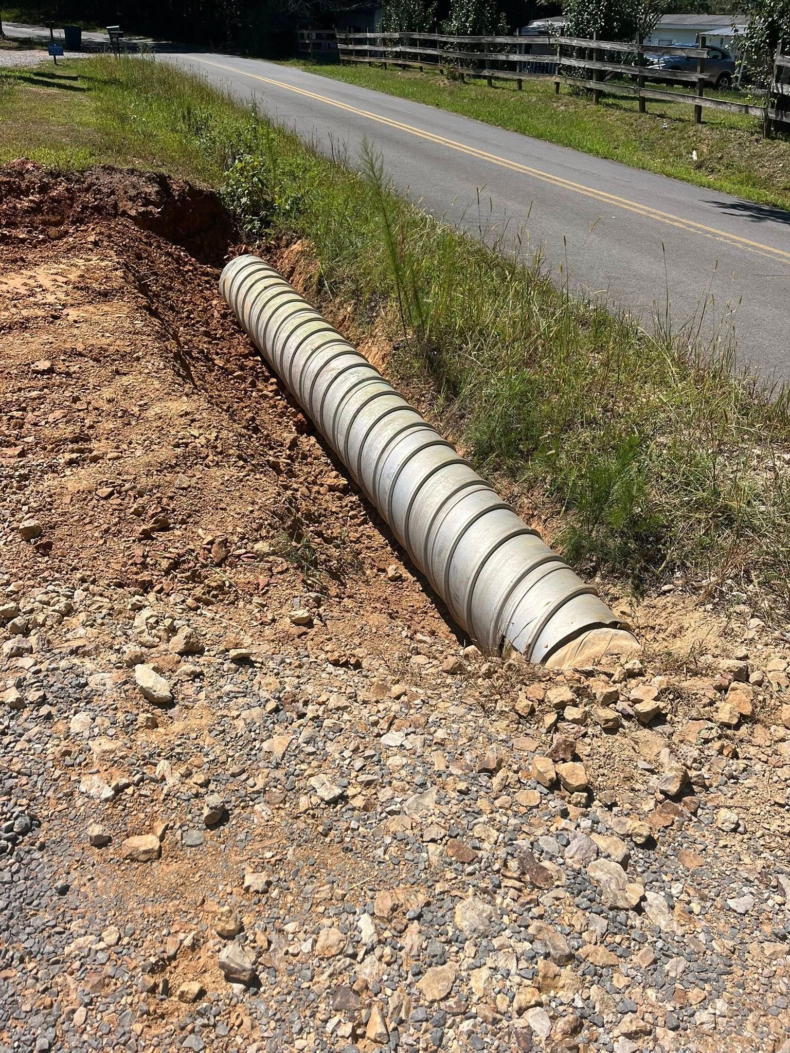 Concrete culvert pipe beside a rural road, with gravel shoulder and green vegetation.