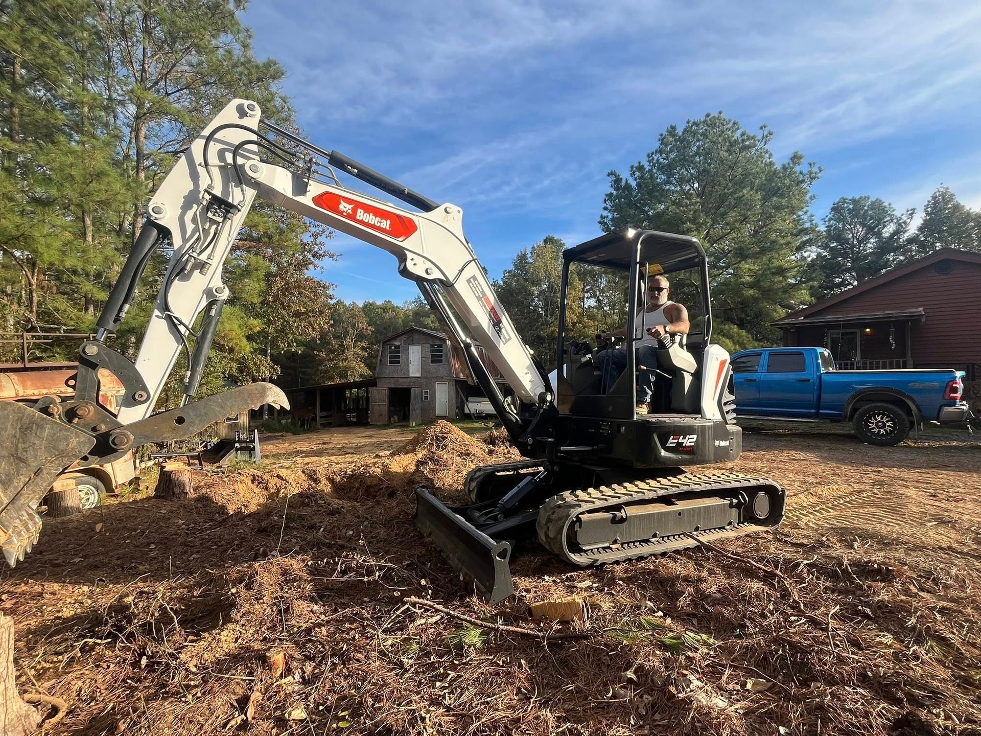 Small excavator clearing dirt at a wooded construction site, with a blue pickup nearby.