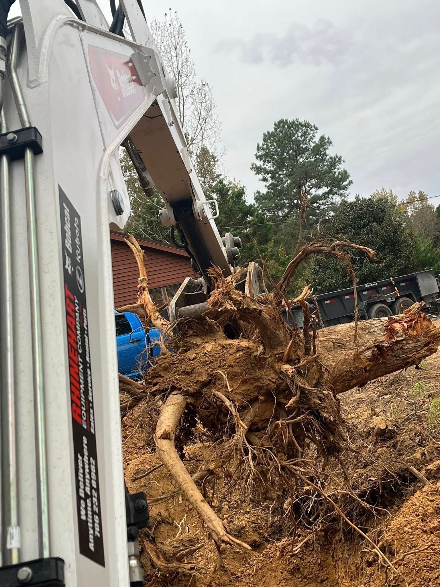 Overturned truck spilling mud and uprooted tree debris beside a house and pine trees