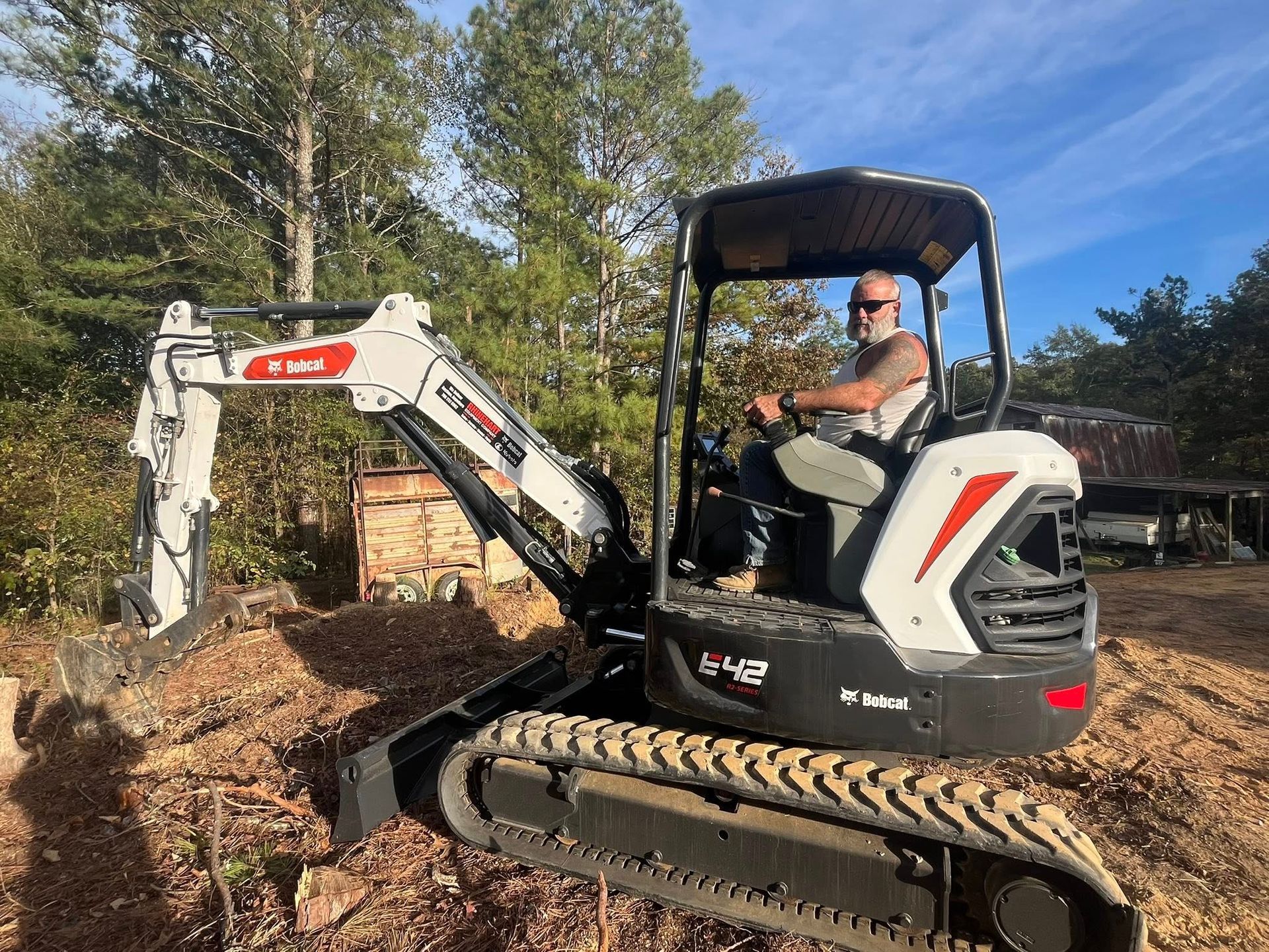 Mini excavator digging in a wooded yard, with an operator in the cab under a blue sky.