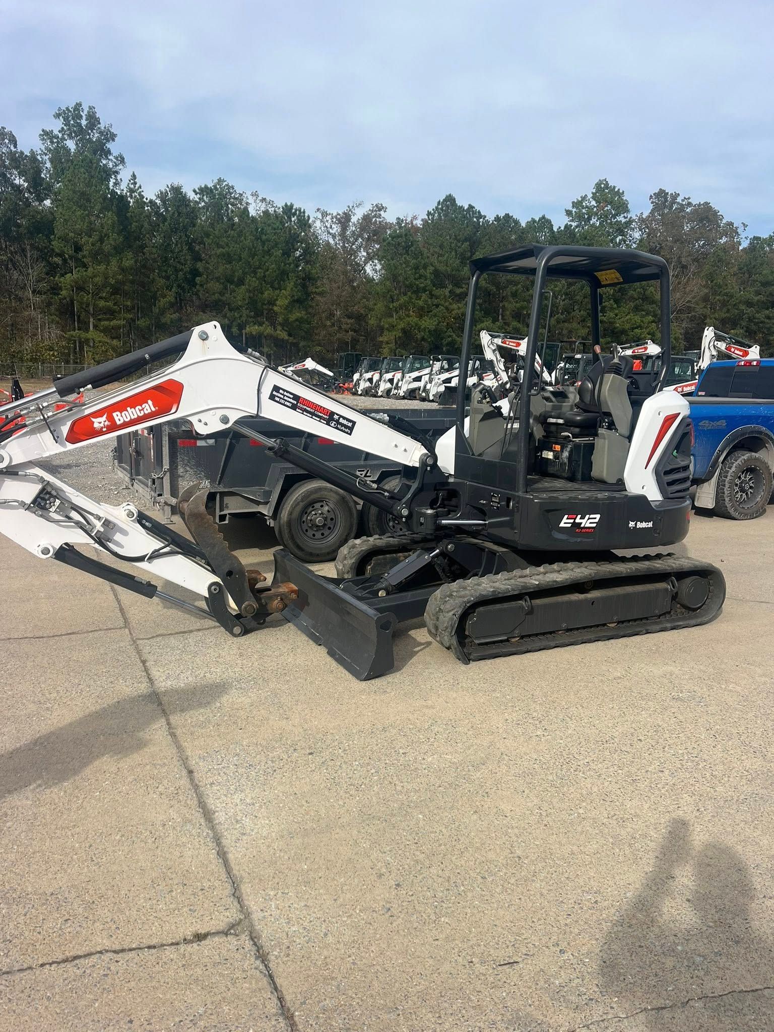 Compact Bobcat excavator with auger attachment parked on pavement outdoors, trees in background