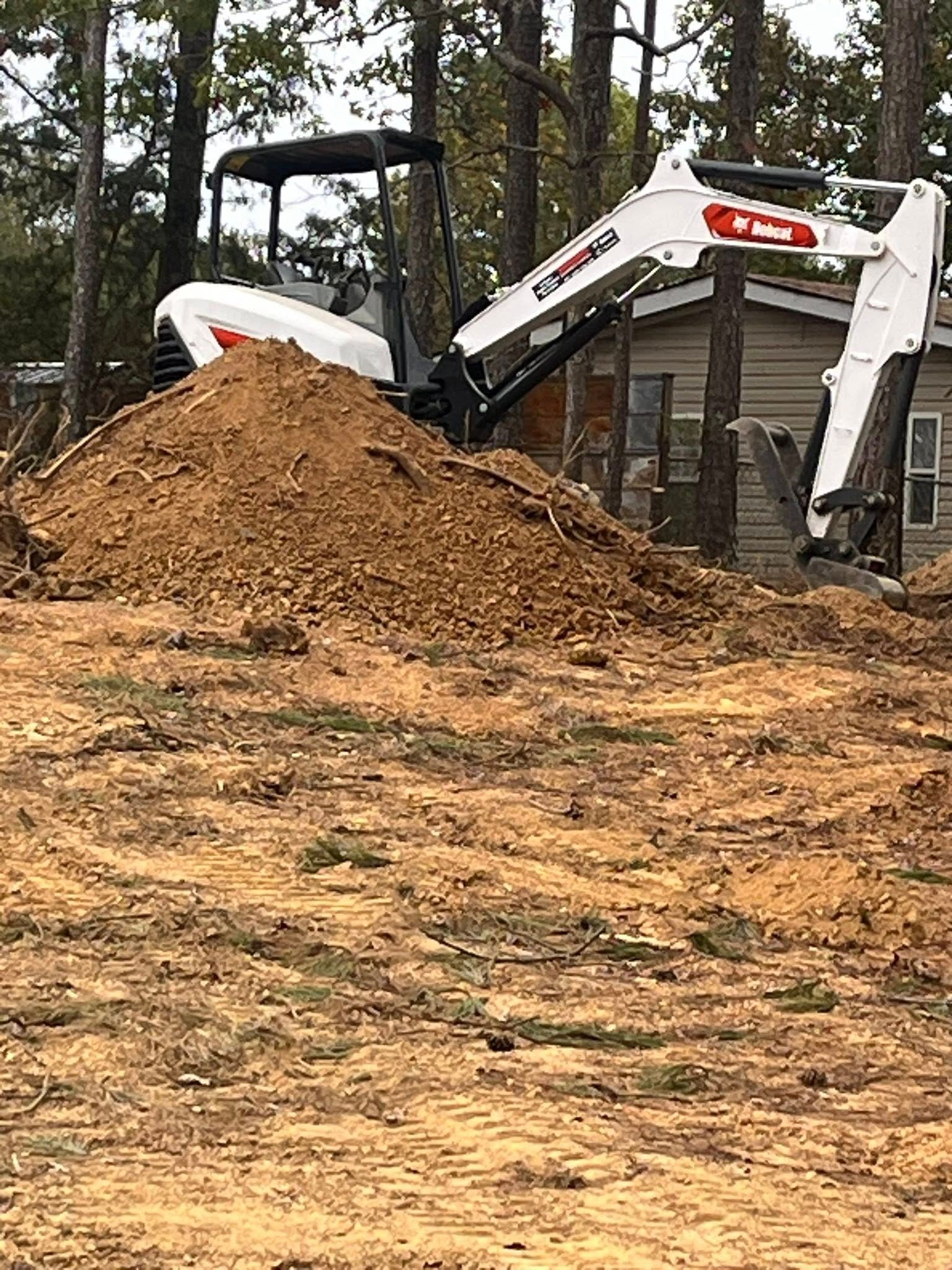 White excavator beside a large dirt pile at a wooded construction site