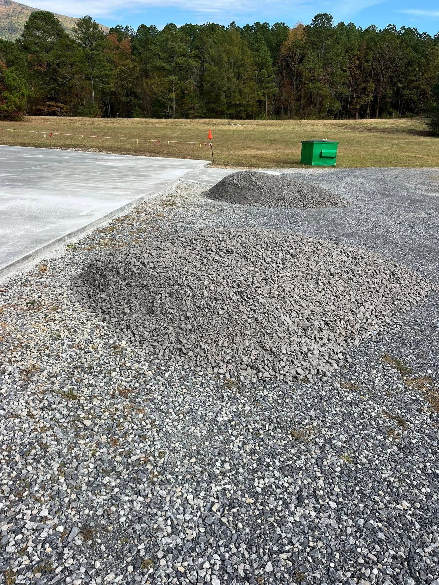Gravel driveway beside a concrete slab, with a green container near a wooded tree line.