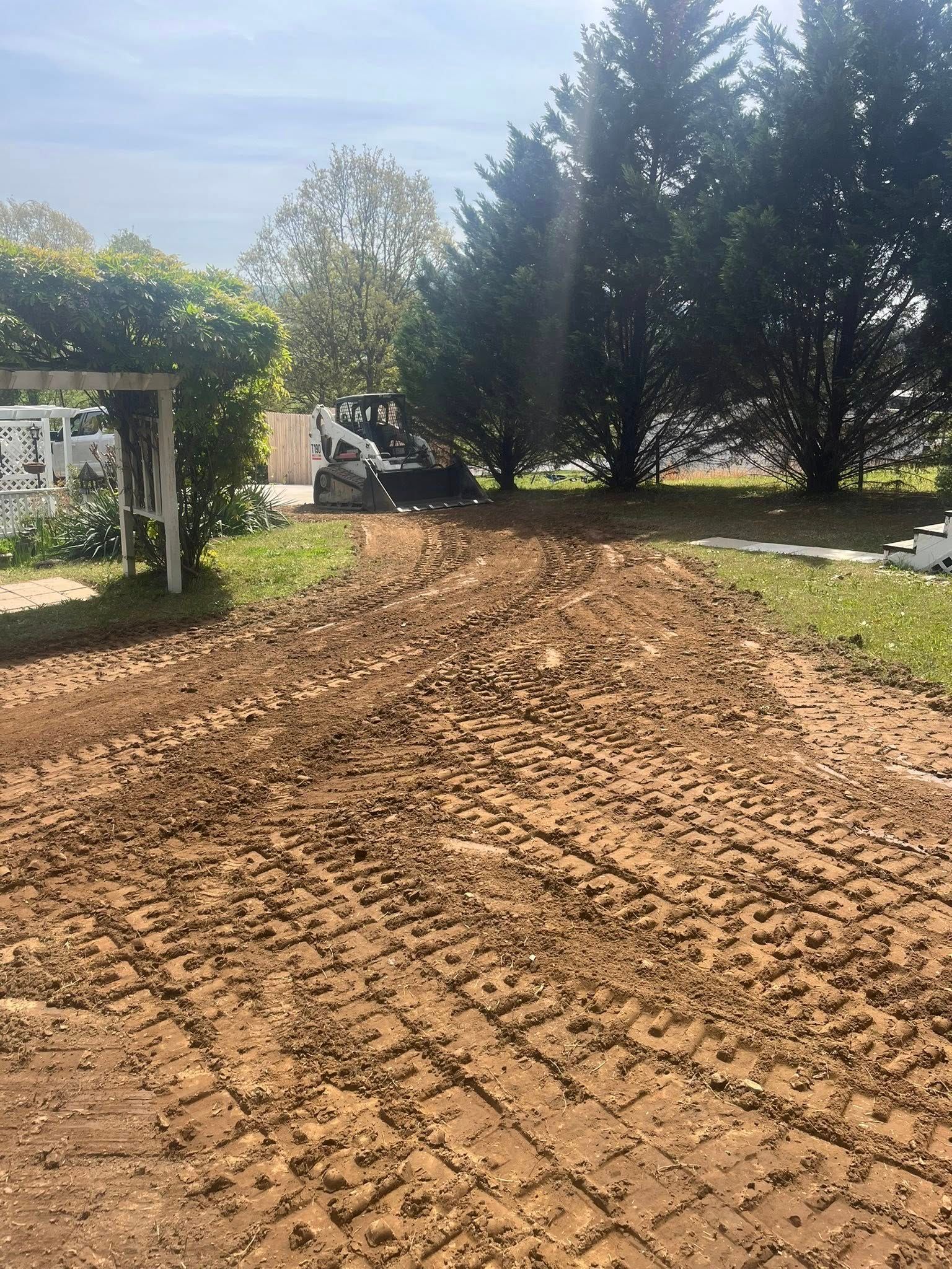 Freshly tilled dirt path in a garden, bordered by grass and trees near a white pergola.