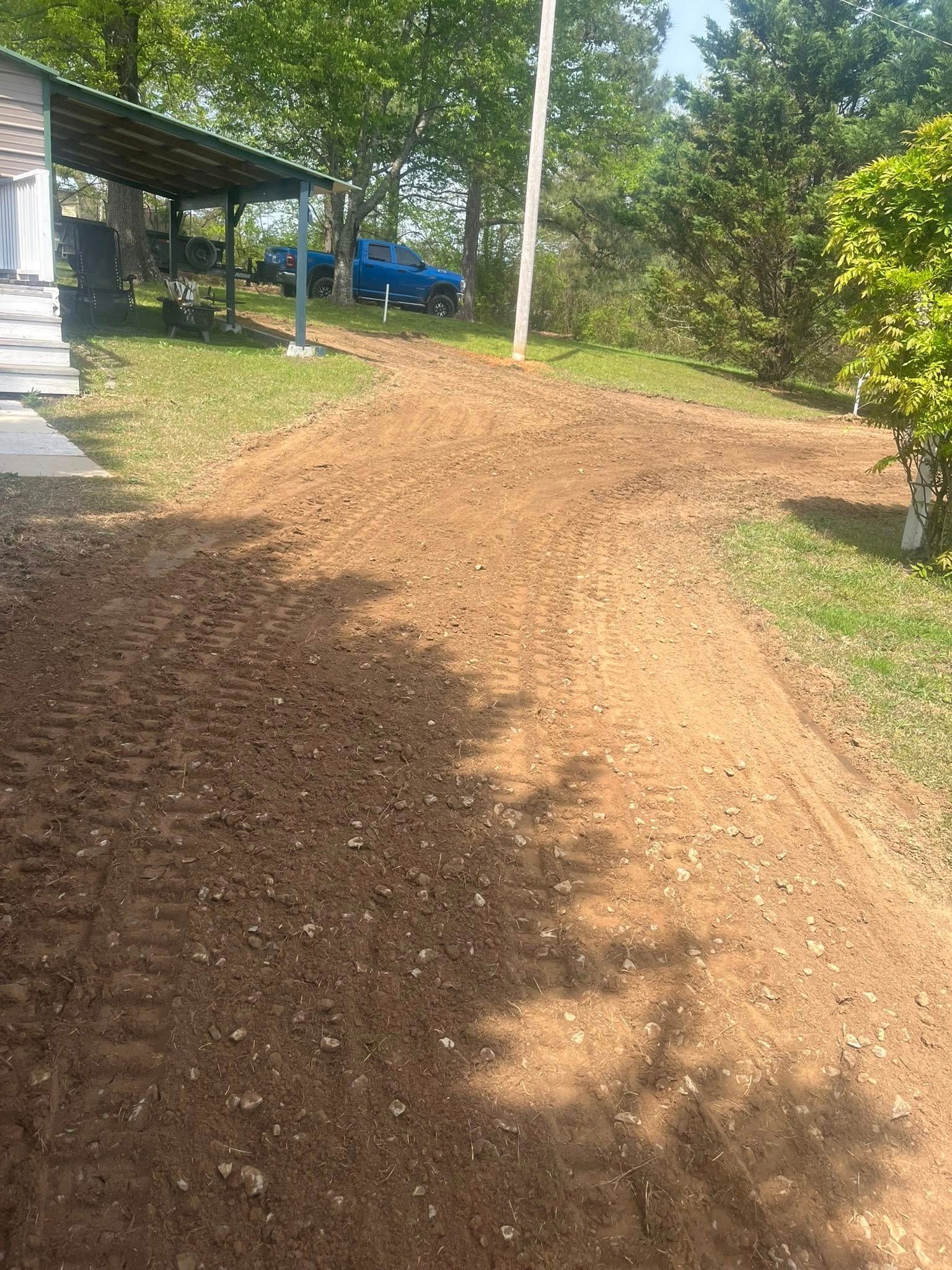Dirt path beside a house and carport, with trees and sunlit yard on either side.