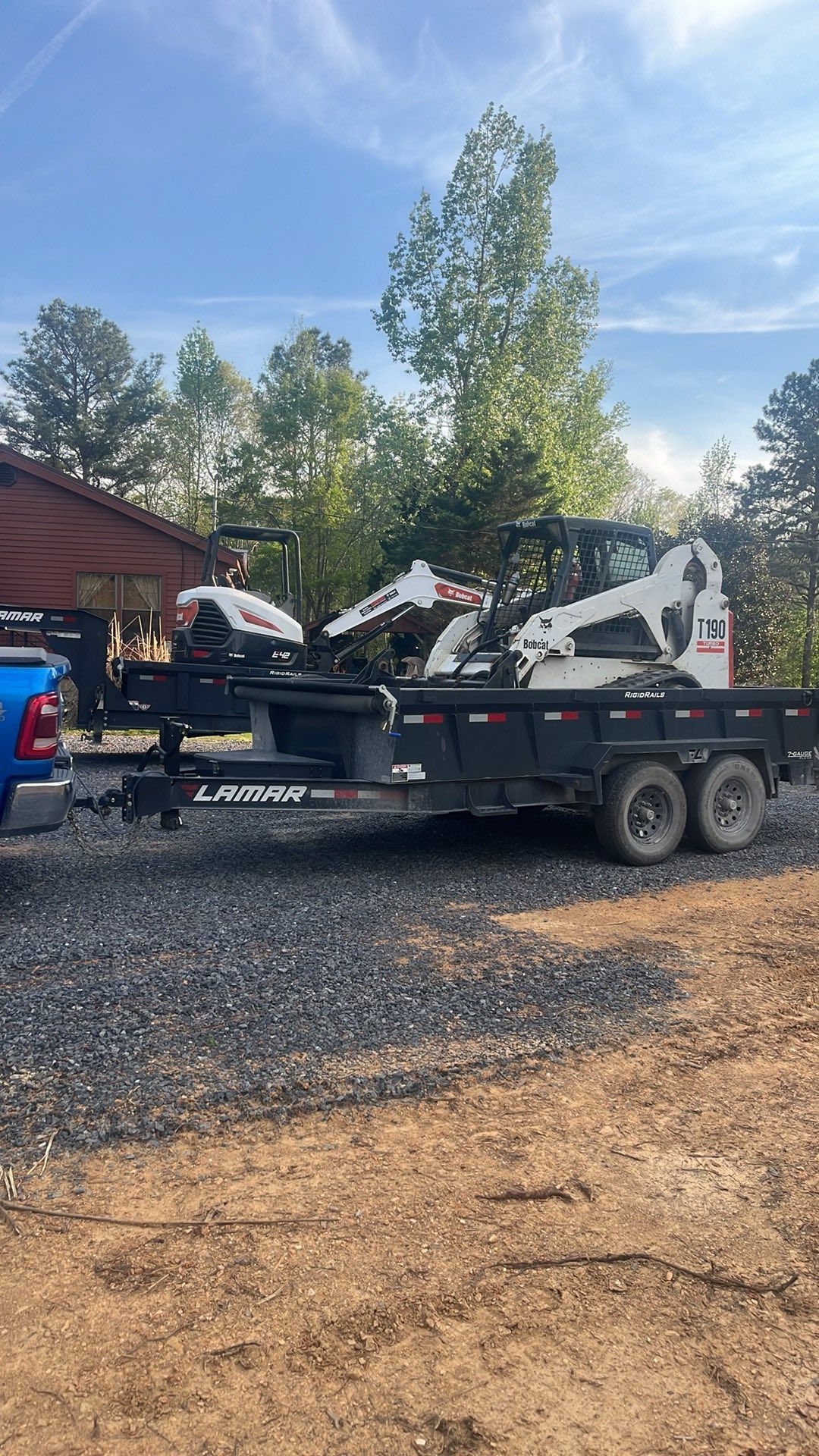 Black trailer loaded with white mini excavators on gravel, parked outdoors near trees and a red building