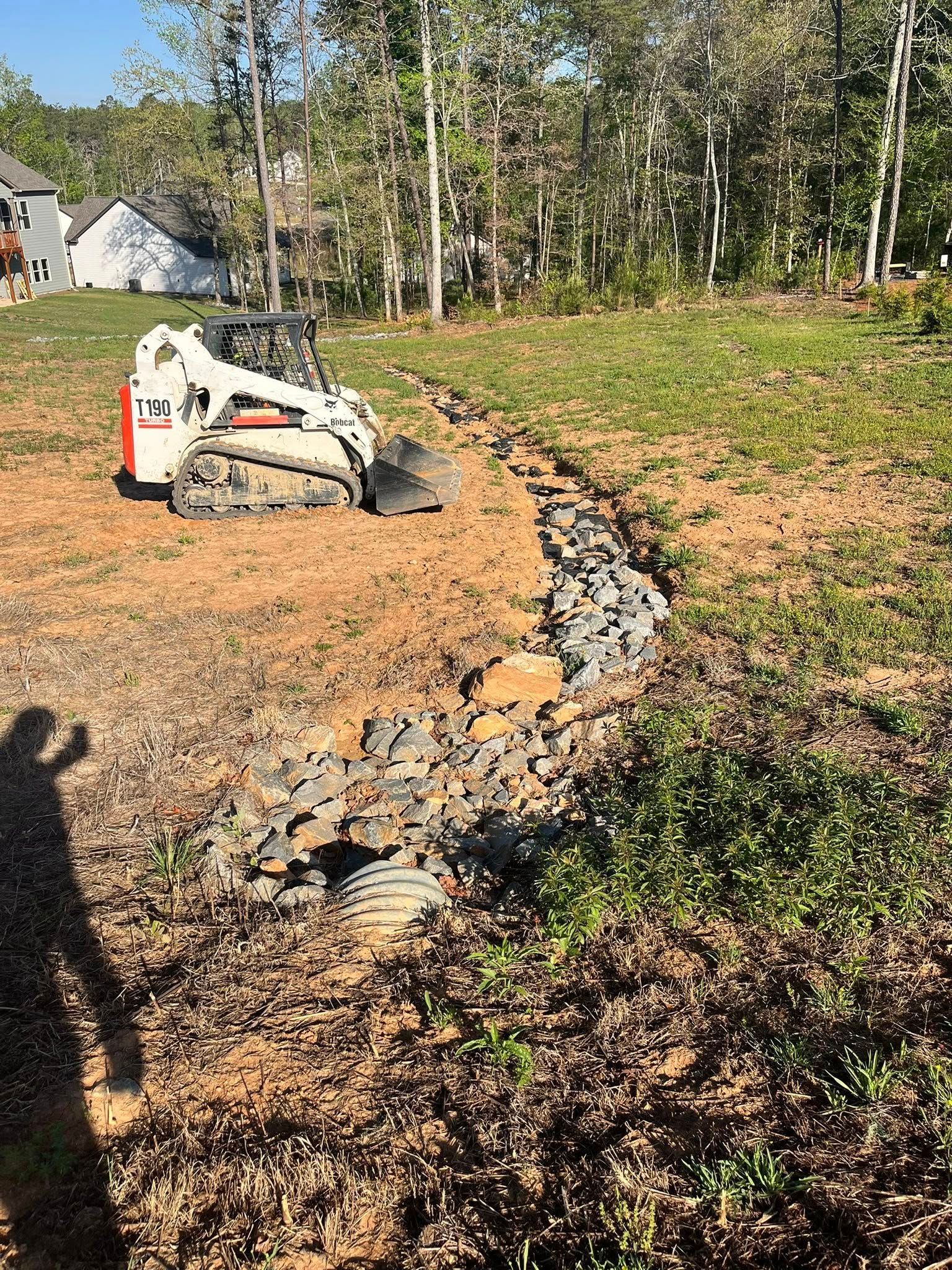 Skid steer placing rocks to form a curved border along a dirt path in a wooded yard