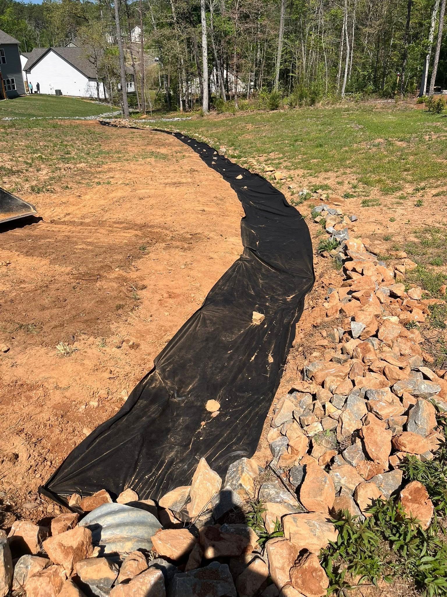 Black silt fence lining a dirt slope beside a rocky retaining edge, with trees and a house in the background