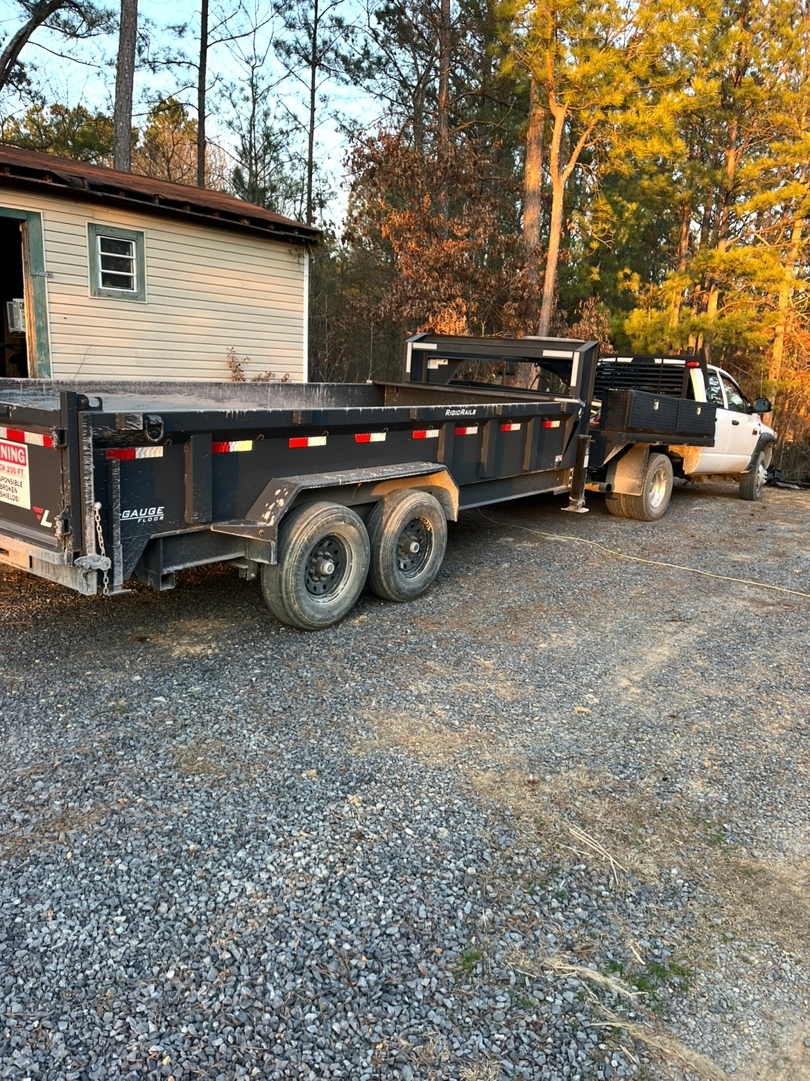 Black dump truck towing a flatbed trailer on a gravel driveway beside a house in autumn.