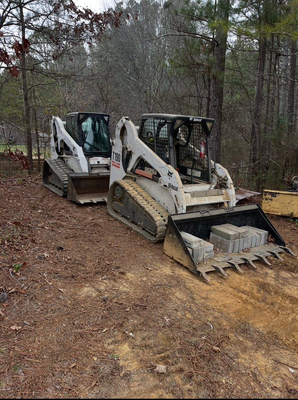 Two white skid-steer loaders parked on a wooded dirt lot, one with a bucket and one with fork attachment