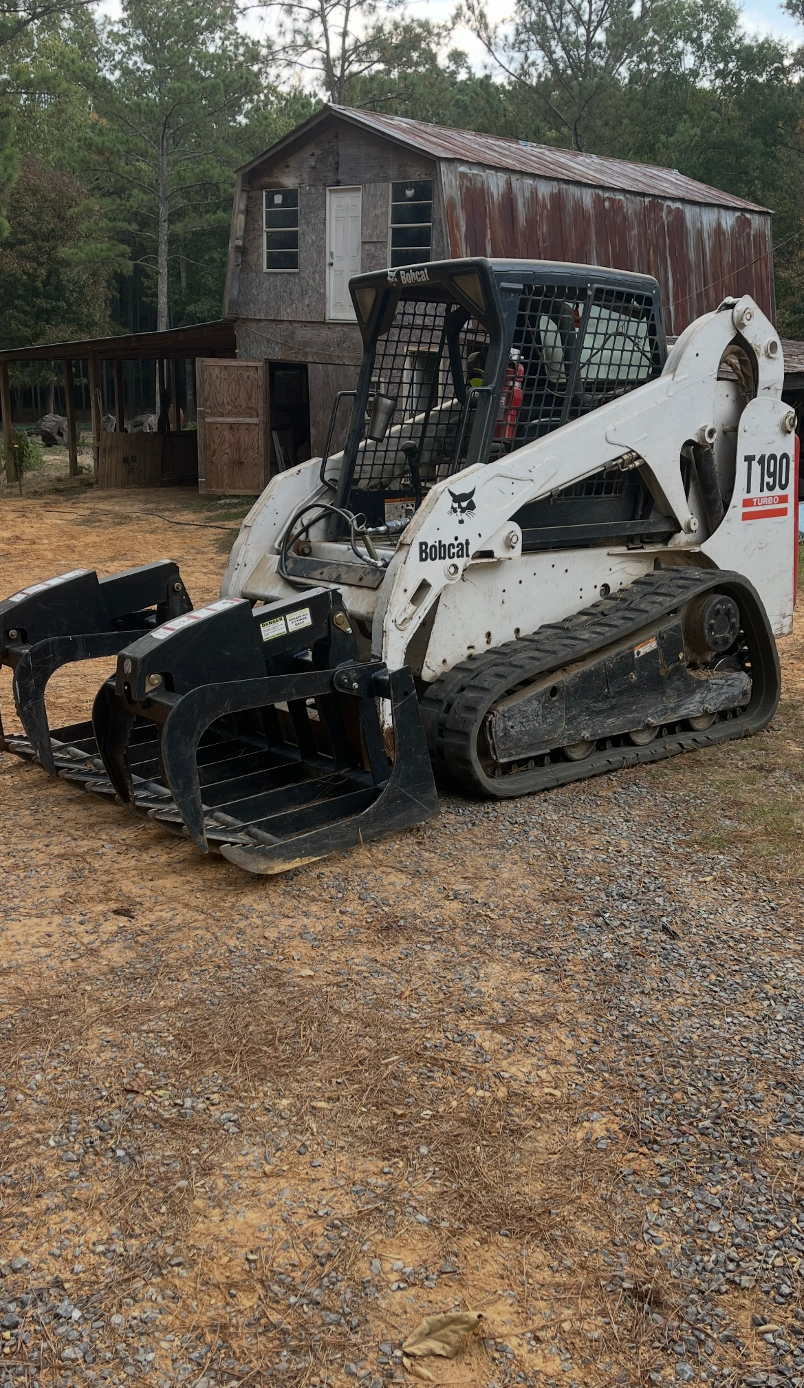 White Bobcat skid steer with a grapple bucket on gravel in front of a weathered barn and trees