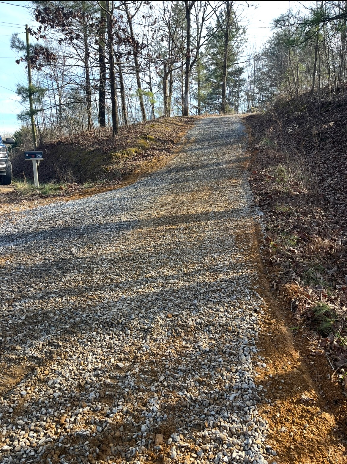 Gravel driveway curving uphill through a wooded area in winter, with bare trees and a mailbox on the left