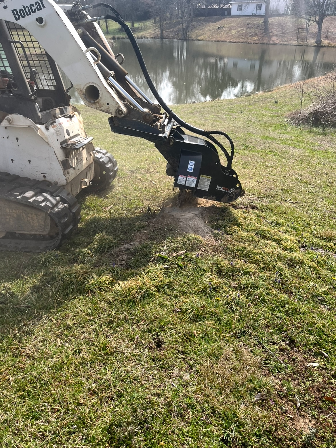 Excavator attachment clearing grass beside a pond on a sunny day