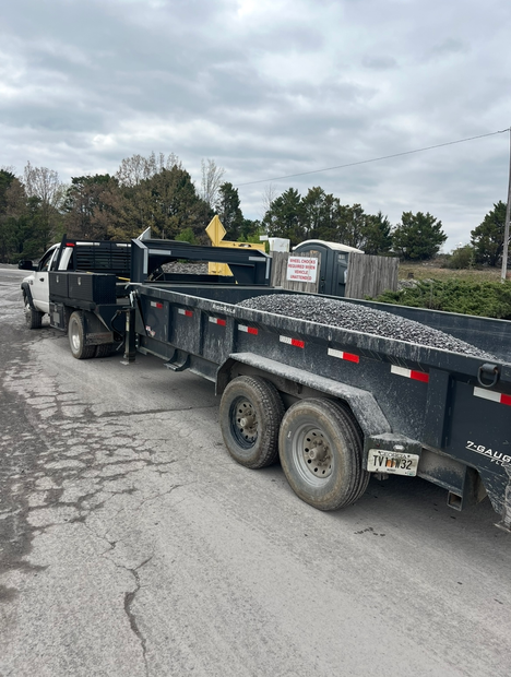 Black pickup truck towing a flatbed trailer loaded with gravel on a roadside under cloudy skies