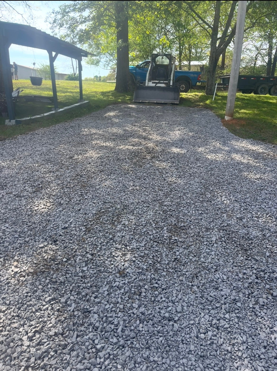 Gravel driveway leading to a tree-covered yard with a vehicle and small structures in the background