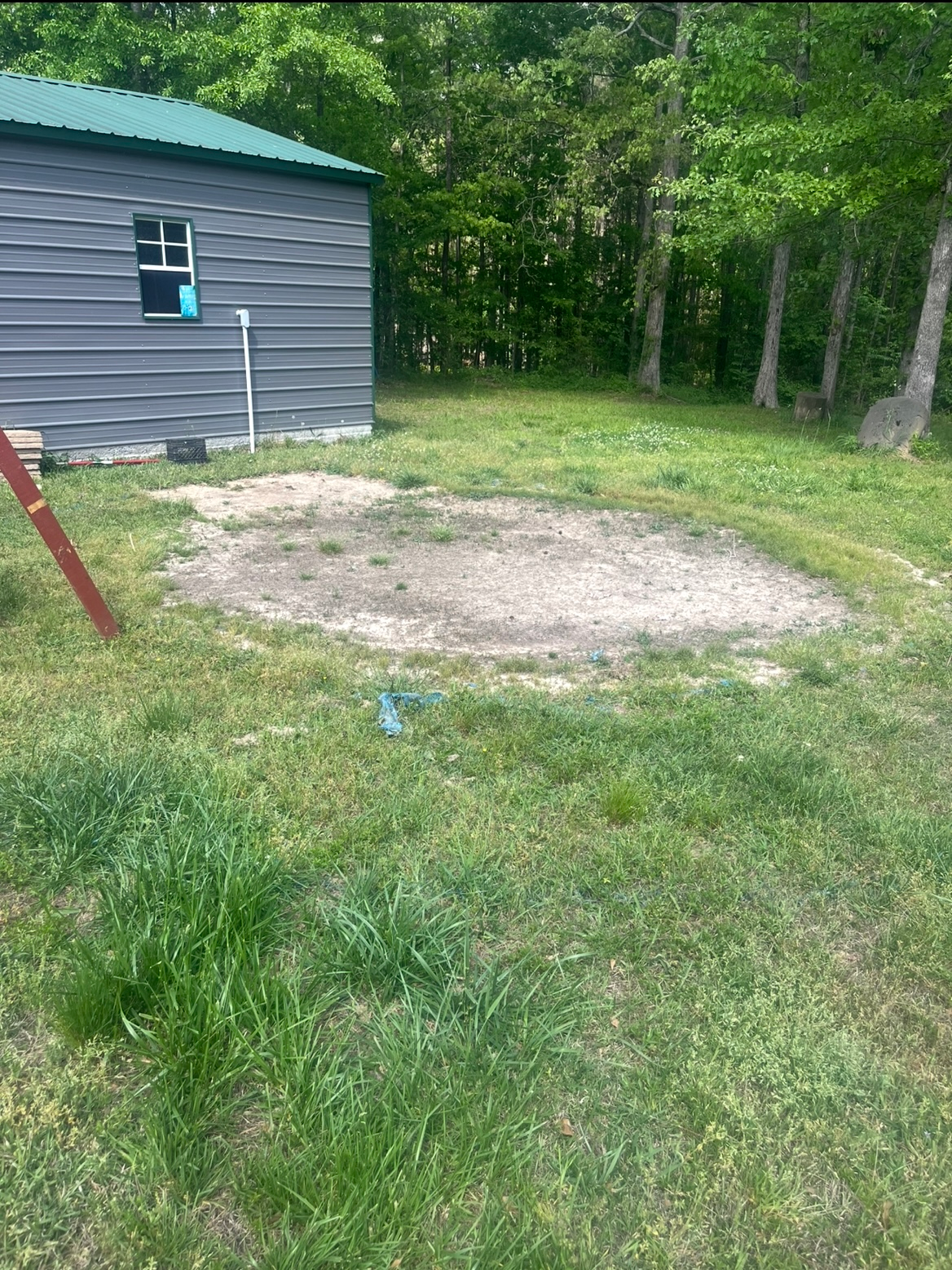 Gravel clearing beside a gray shed in a wooded yard, with grass and trees around it