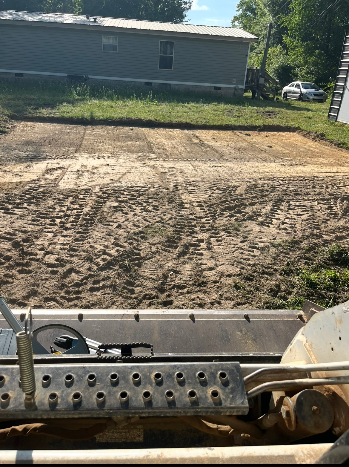 Construction equipment leveling a dirt lot beside a mobile home and parked car.