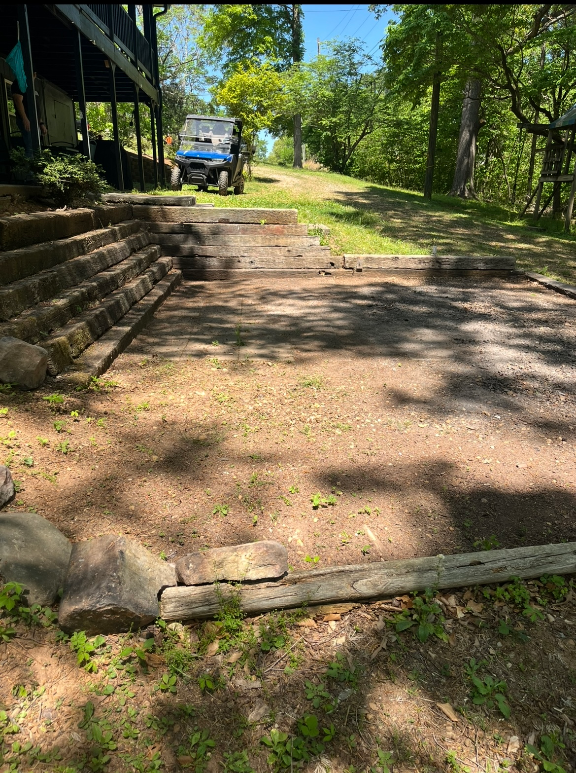 Gravel driveway beside a wooden porch, leading uphill past trees with a blue vehicle in the background