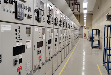 Rows of industrial electrical control panels in a bright utility corridor with a yellow floor line