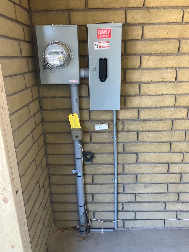 Electrical meter and service panel mounted on a brick wall with exposed conduit and piping.