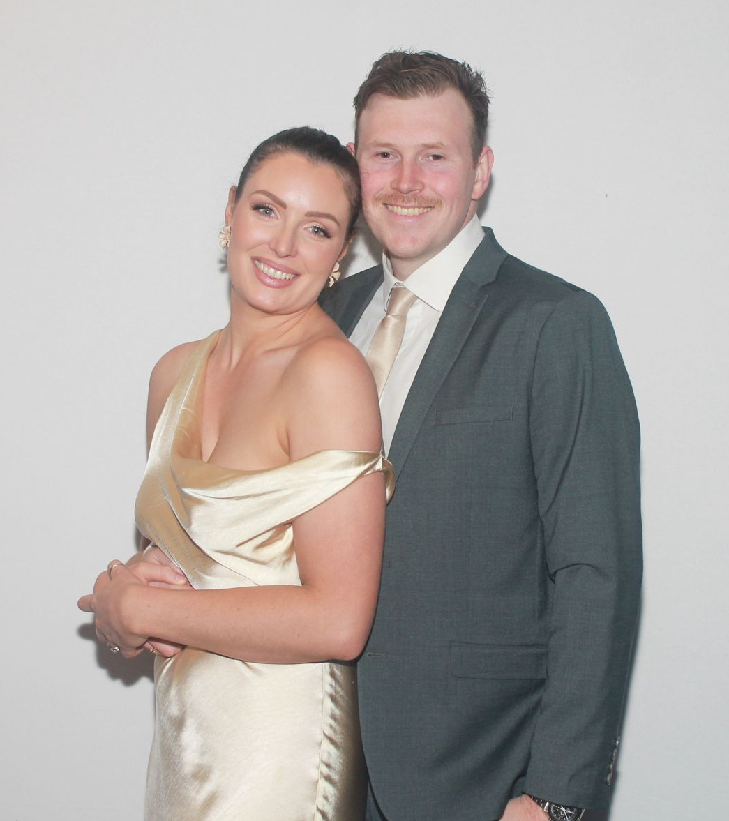 A smiling couple posing together, the woman in a satin champagne-colored gown and the man in a charcoal suit and tie.