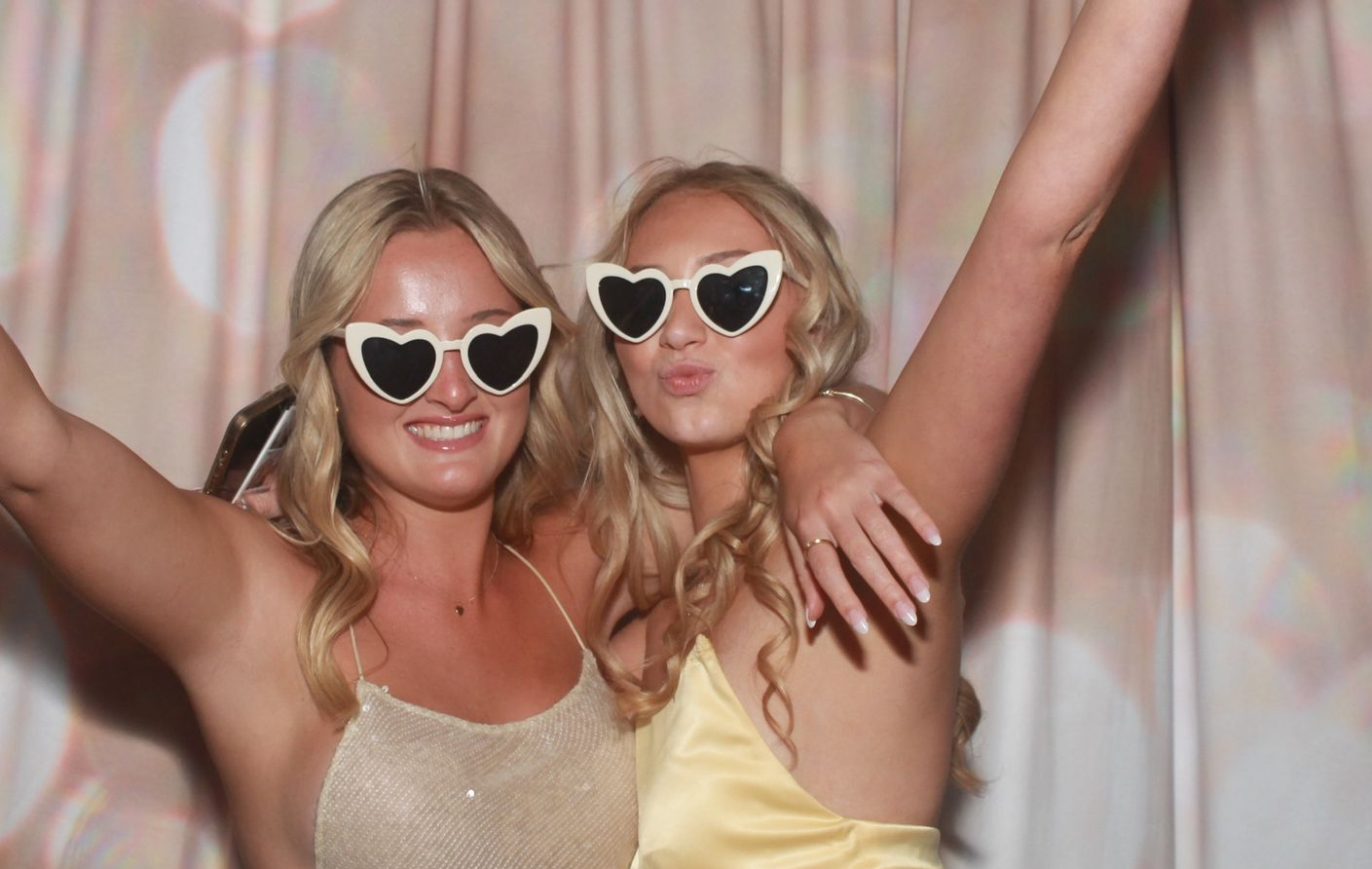 Two young women in heart-shaped sunglasses pose for a photo in front of a shimmery light-colored curtain.