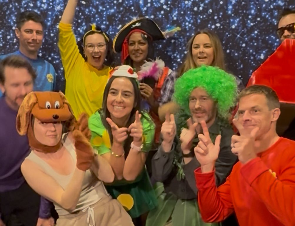 Group of people in costumes, posing in front of a starry backdrop; some wear hats/wigs and make hand gestures.