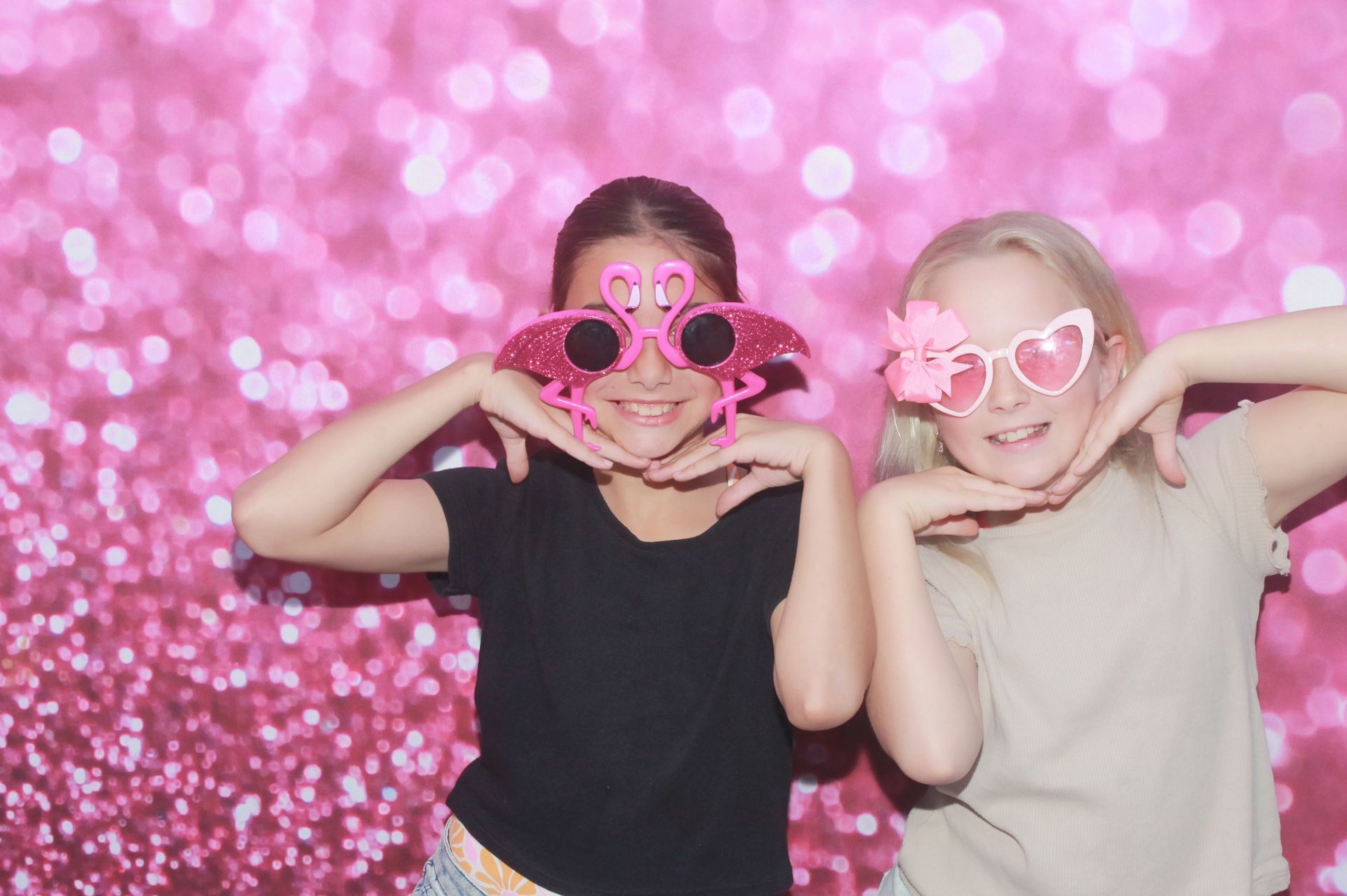Two girls wearing novelty glasses pose in front of a pink glitter backdrop.