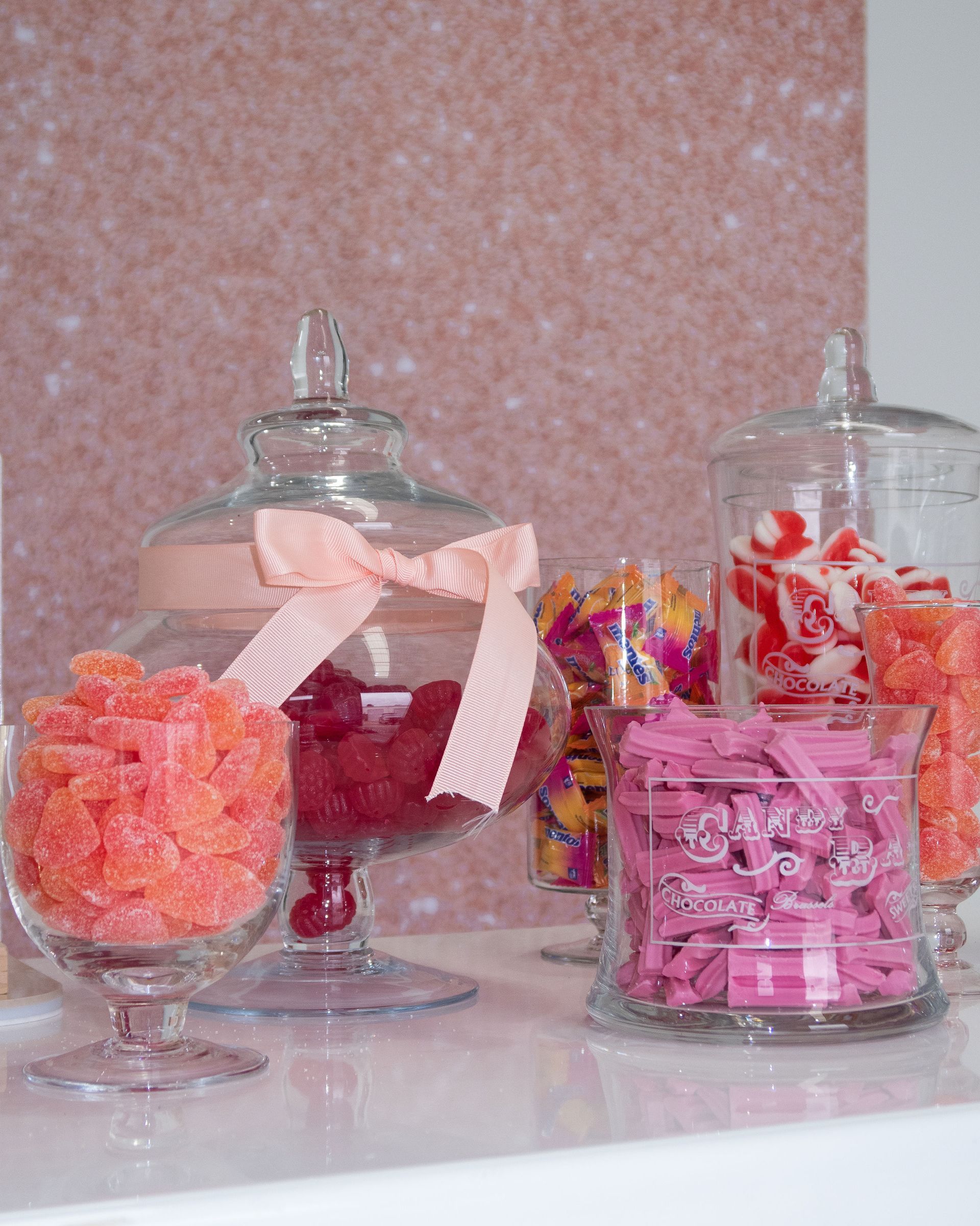 Glass jars filled with pink and orange candy, displayed against a pink glitter background.