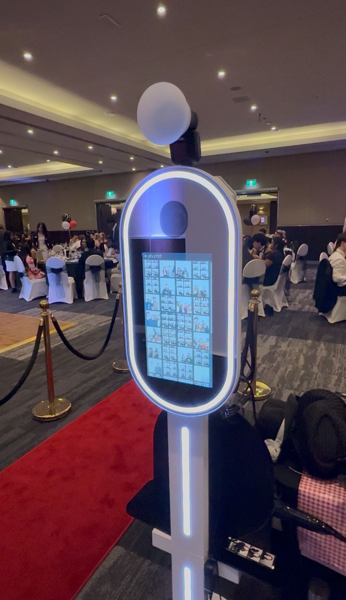 Photo booth at an event with a red carpet and guests in the background. The booth has a glowing outline and screen.