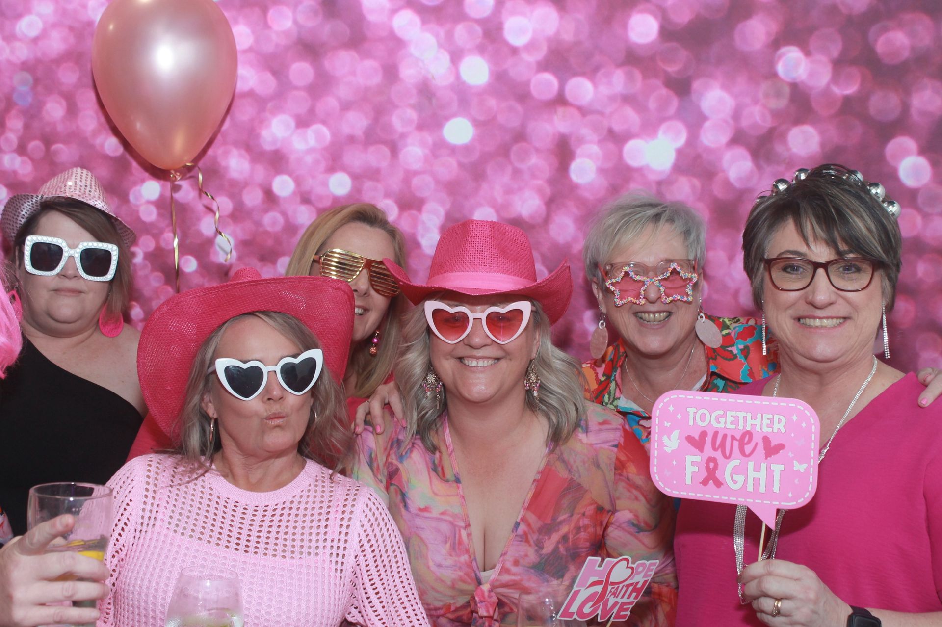 Group of women in pink attire with props, posing in front of a sparkly pink backdrop.