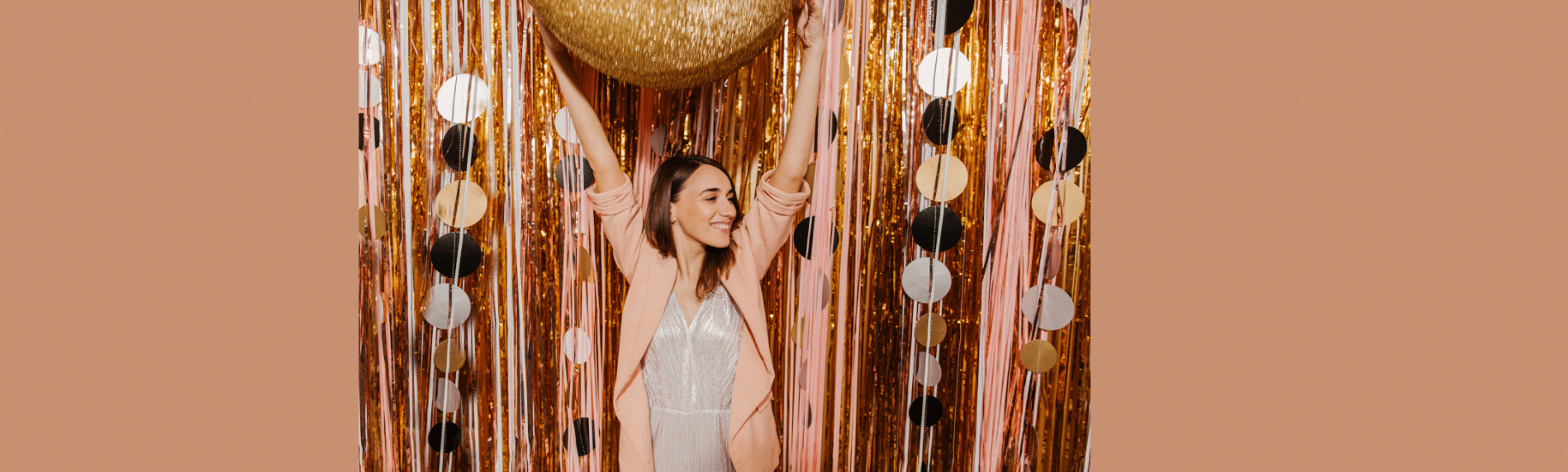 Woman raising arms in front of a gold tinsel backdrop with a large gold disco ball.