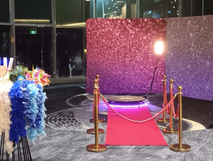Photo booth setup with red carpet, gold stanchions, pink glitter backdrop, and bright spotlight.