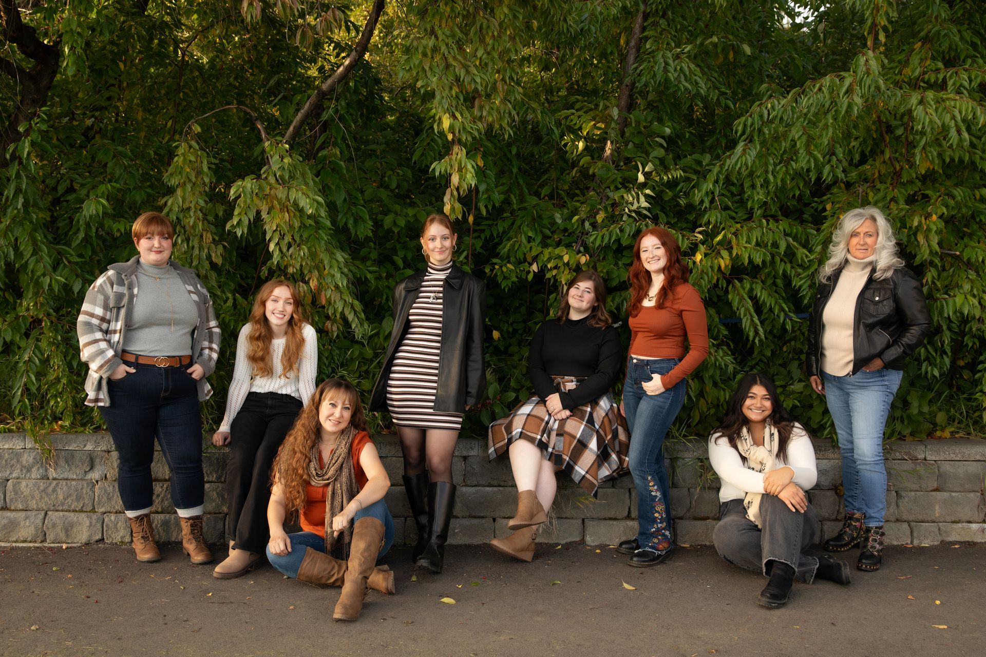 Group of eight women posing outdoors in front of green foliage. They are dressed in fall attire.