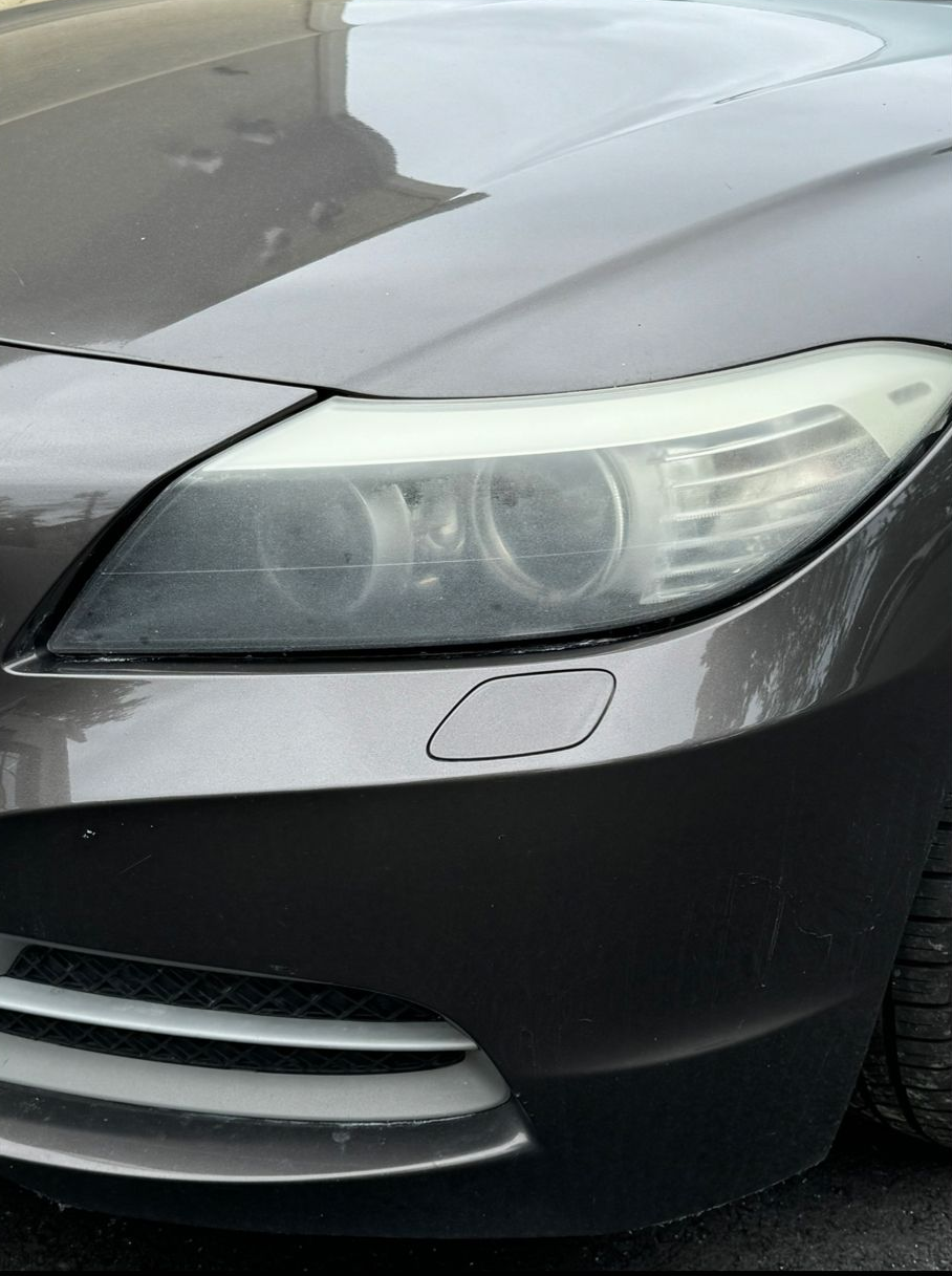 A close-up of a grey car's front bumper, featuring a headlight, a headlight washer cap, and part of the lower grille.