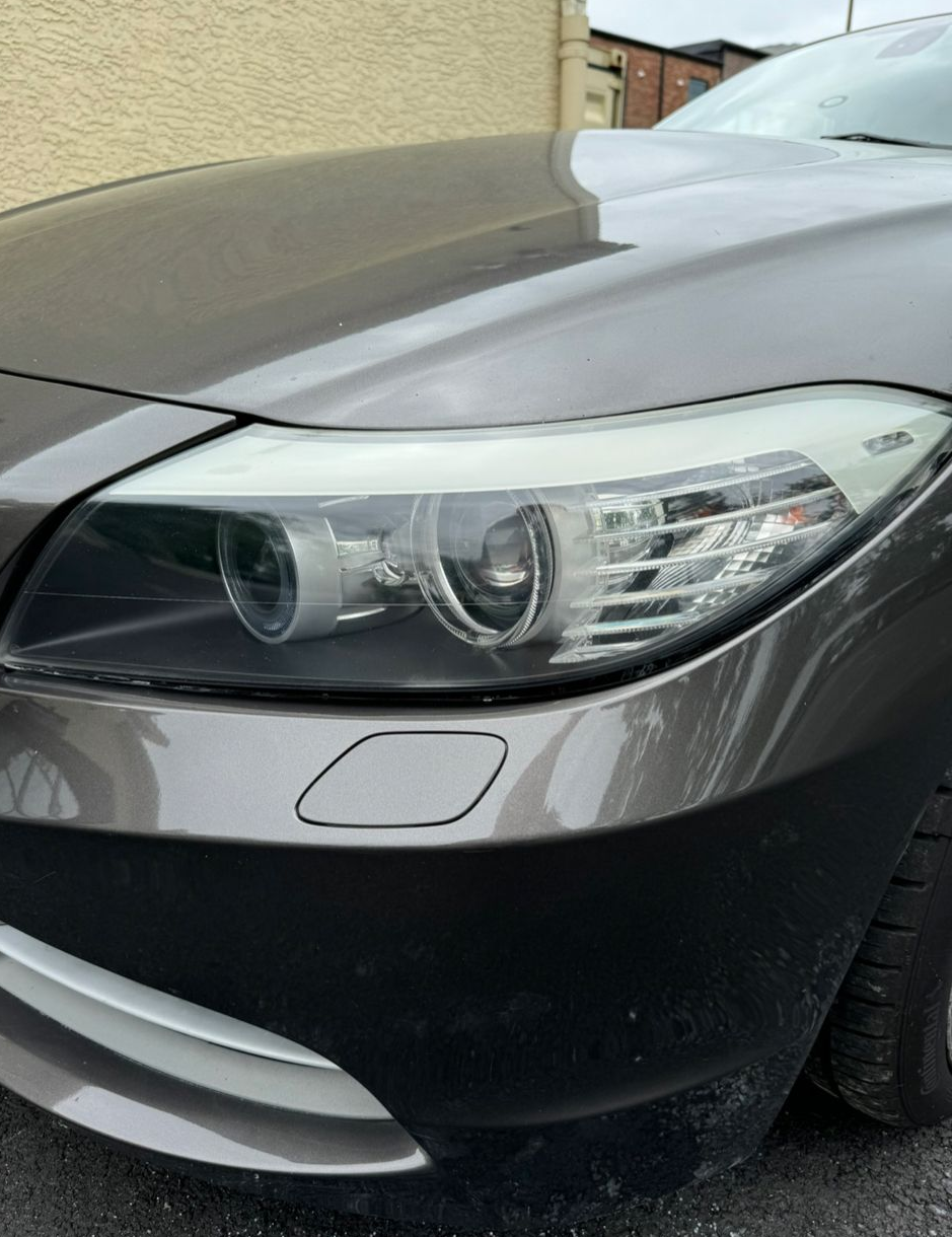 A close-up view of the front passenger-side headlight and bumper of a metallic brown sedan parked outdoors.