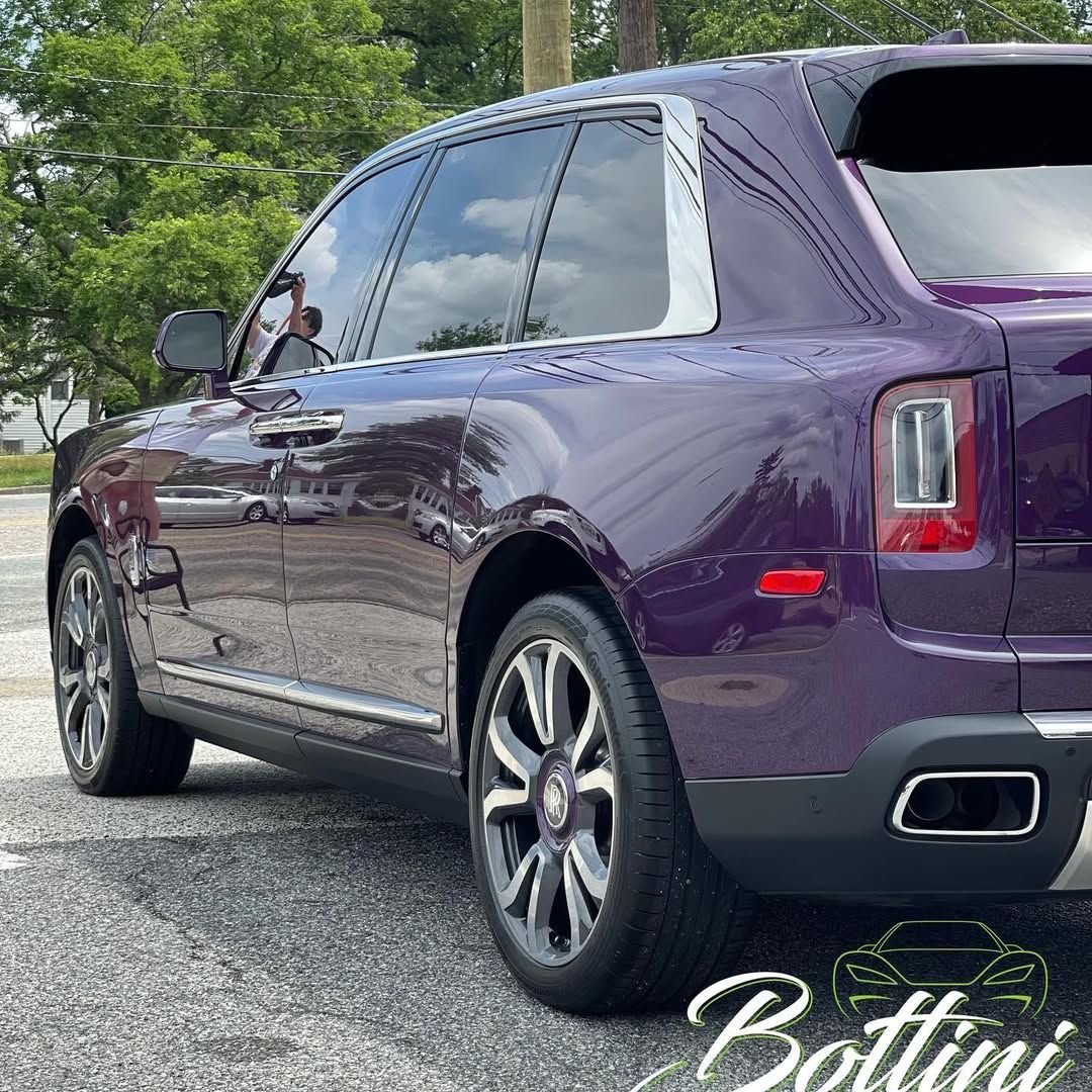 A deep purple Rolls-Royce Cullinan SUV parked on pavement with a logo watermark in the bottom right corner.