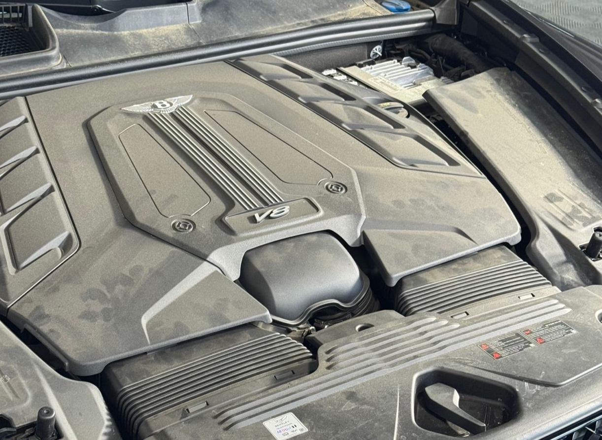A dusty, V8 engine bay featuring a black cover with the Bentley logo, housed inside a vehicle's engine compartment.