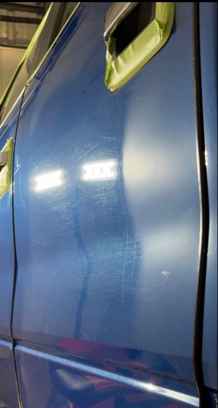 A side view of a blue car door with taped handles showing swirl marks on the paint surface under bright lighting.