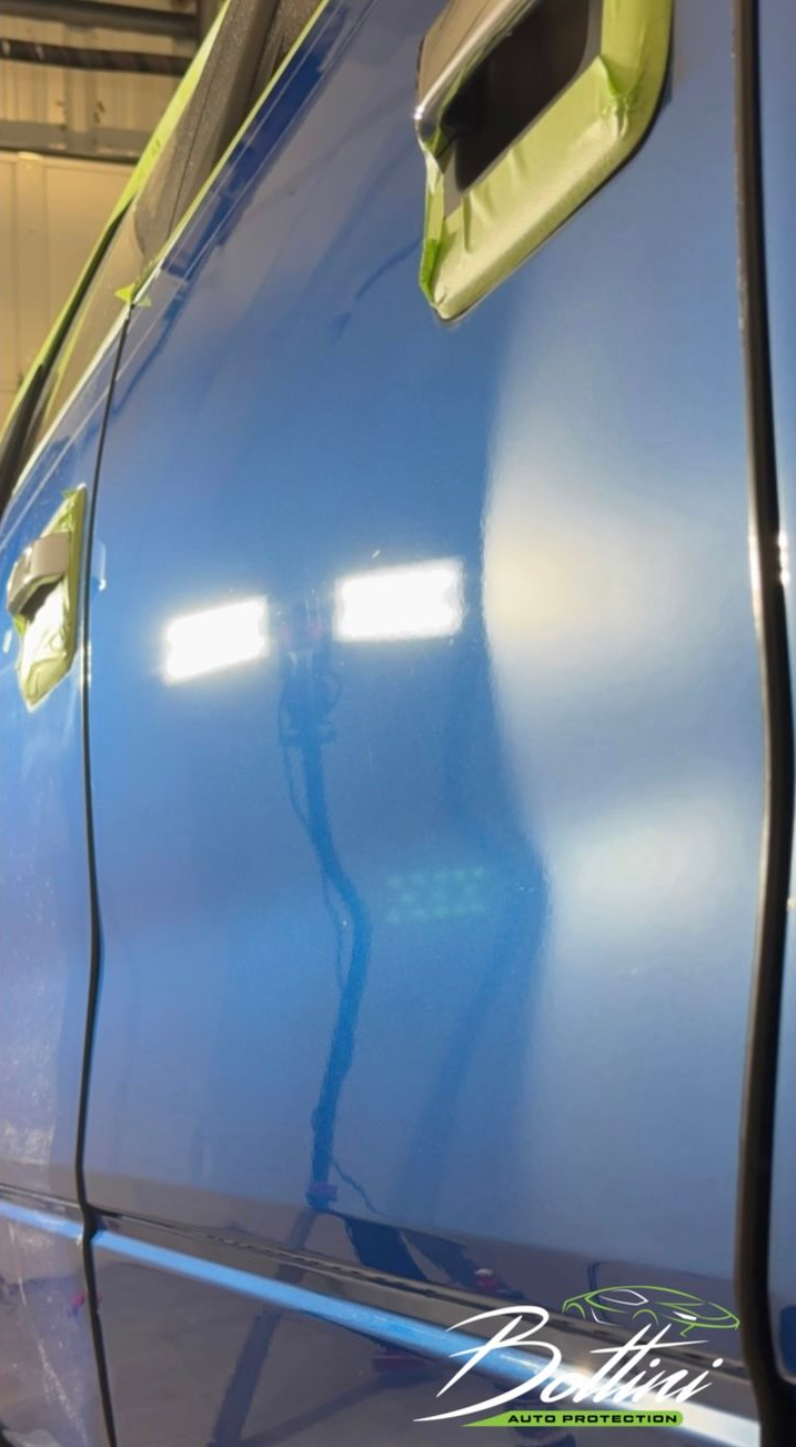Bright blue car door with masking tape on the handle, showing a reflection of shop lights.