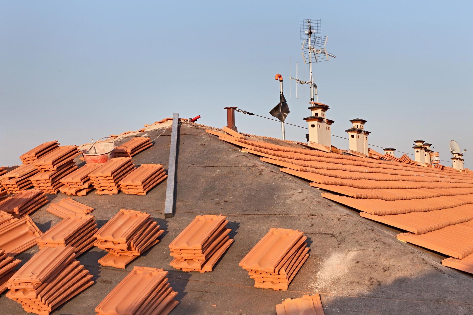 Un tetto residenziale in riparazione, con pile di tegole di terracotta e una falda parzialmente coperta, sotto un cielo limpido.