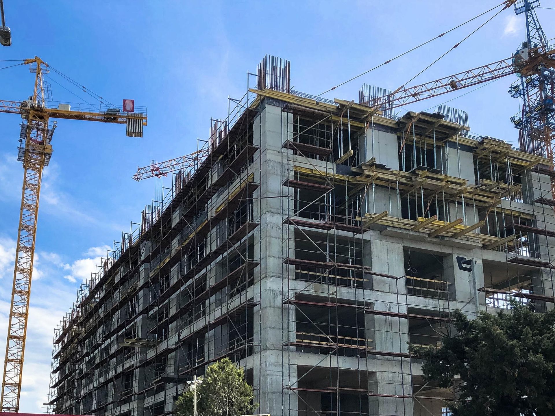 Un edificio in cemento armato a più piani in costruzione, con impalcature, circondato da alte gru gialle contro un cielo azzurro.