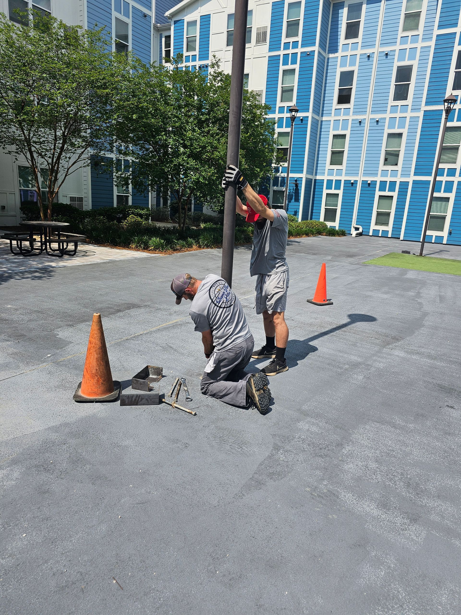 Two men are working on a pole in a parking lot in front of a blue building - Milton, FL - Damn Fine Jobs