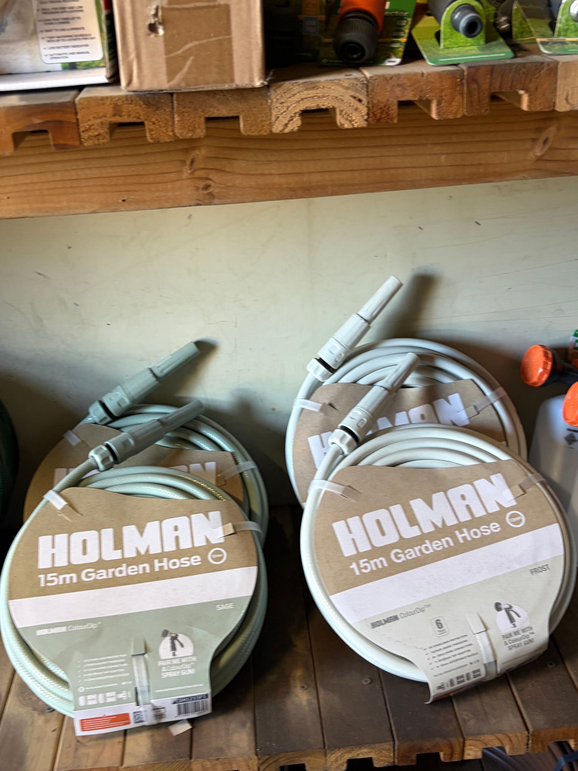 Two coiled HOLMAN garden hoses on a warehouse shelf, one green and one white in cardboard packaging