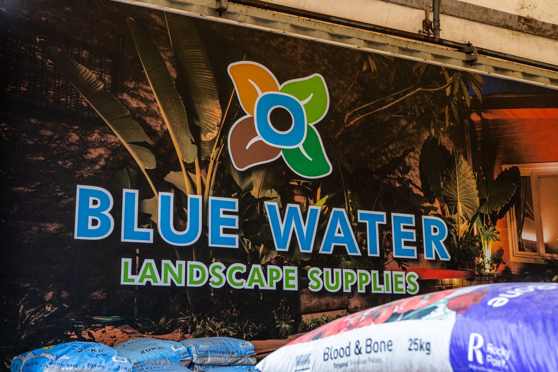 Blue Water Landscape Supplies sign with a colorful flower logo above stacked garden materials.
