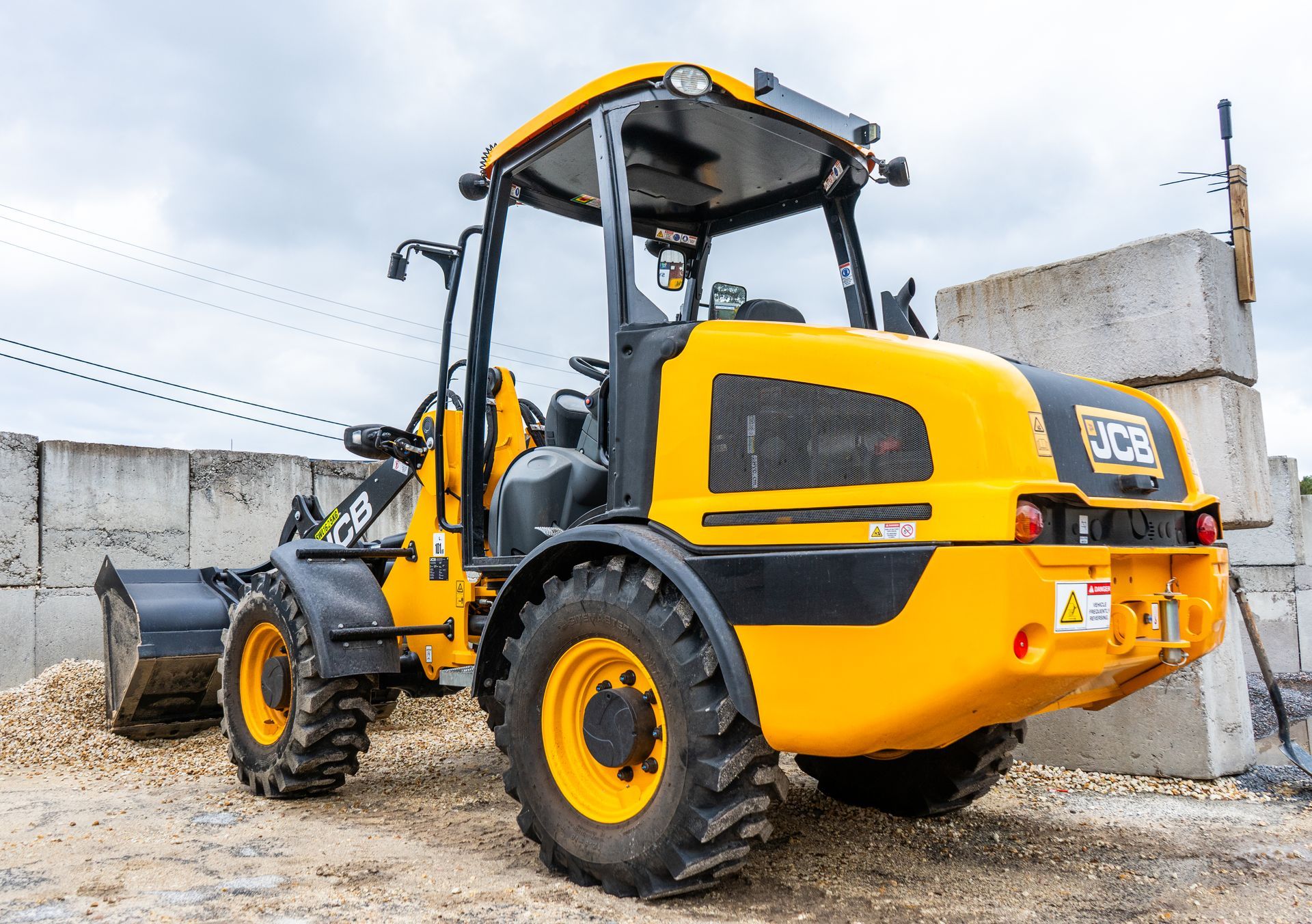 Yellow backhoe loader parked on a gravel lot beside concrete walls under a cloudy sky